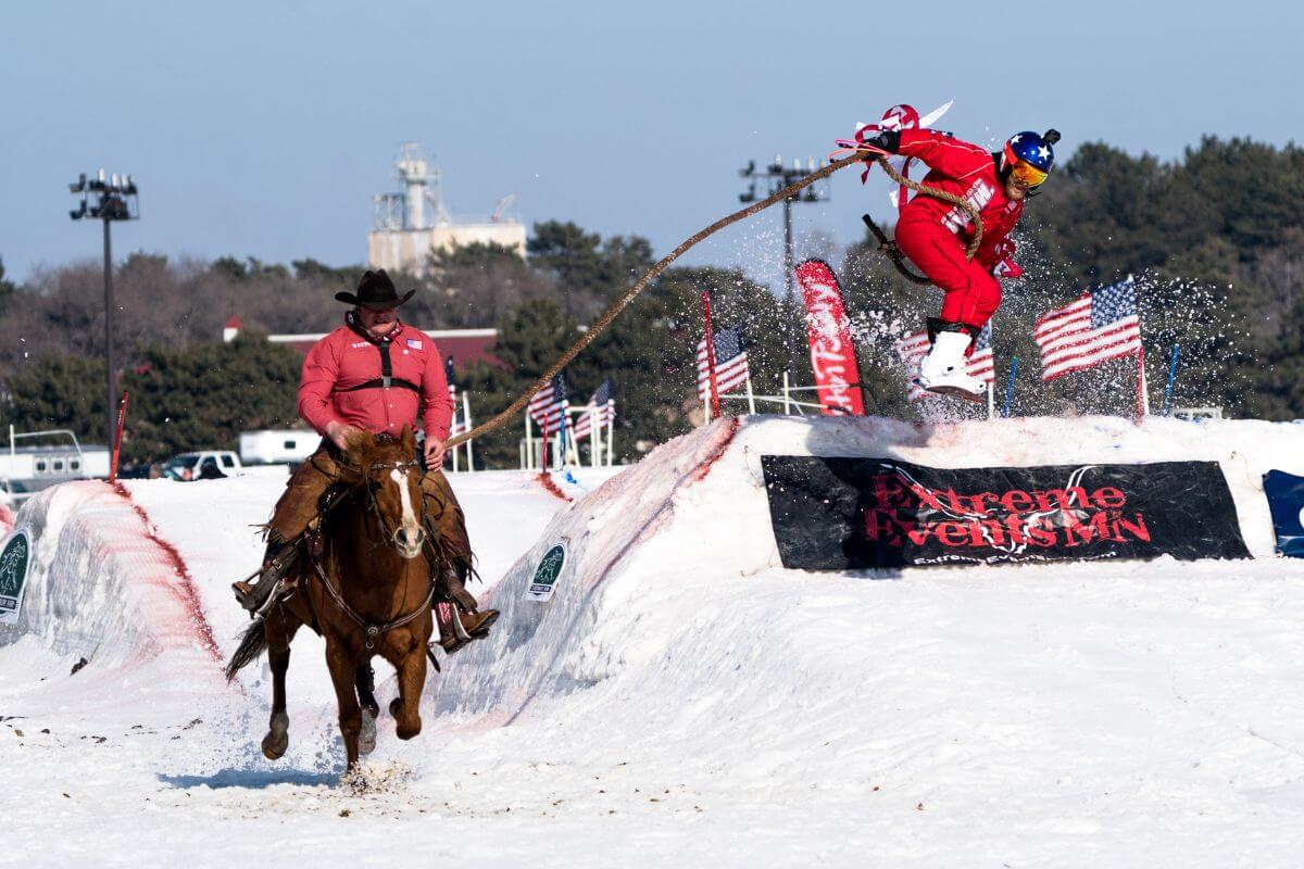 Two men at a skijoring competition in Montana, one on horseback and the other rider jumping over obstacles with a rope.