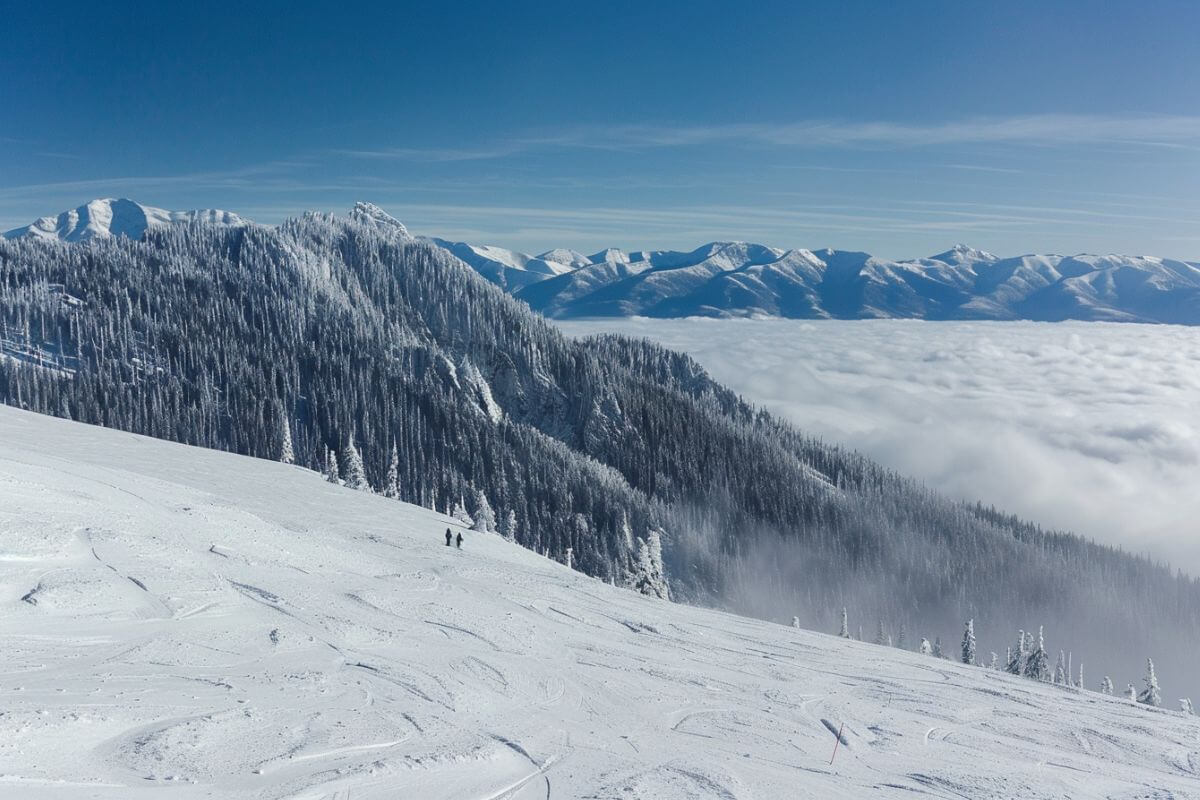 Two skiers are skiing down a snowy slope in Whitefish Mountain Resort with clouds, mountains and trees in the background.