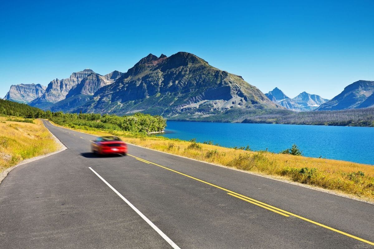 A red car driving down a road next to a lake with mountains in the background in Montana.
